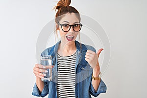 Young beautiful redhead woman drinking glass of water over isolated white background pointing and showing with thumb up to the