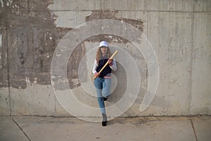 Young and beautiful redhead woman is happy with baseball cap, jacket, baseball bat and jeans, she is posing in front of grey