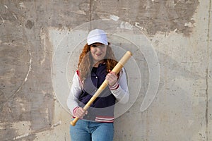 Young and beautiful redhead woman is happy with baseball cap, jacket, baseball bat and jeans, she is posing in front of grey