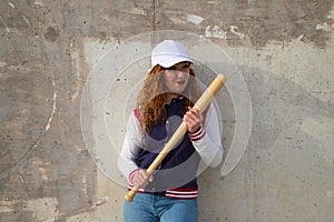 Young and beautiful redhead woman is happy with baseball cap, jacket, baseball bat and jeans, she is posing in front of grey