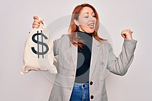 Young beautiful redhead woman holding bag with money and dollar sign over white background pointing and showing with thumb up to