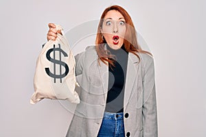 Young beautiful redhead woman holding bag with money and dollar sign over white background scared in shock with a surprise face,