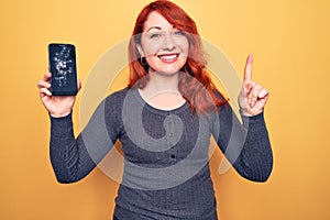 Young beautiful redhead woman holding broken smartphone showing cracked screen smiling with an idea or question pointing finger