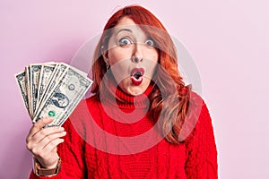 Young beautiful redhead woman holding bunch of dollars banknotes over pink background scared and amazed with open mouth for