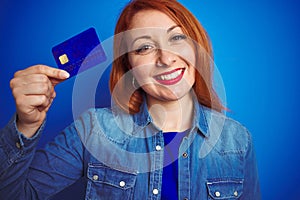 Young beautiful redhead woman holding credit card over blue isolated background with a happy face standing and smiling with a