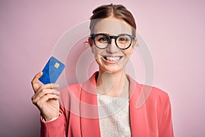 Young beautiful redhead woman holding credit card over isolated pink background with a happy face standing and smiling with a