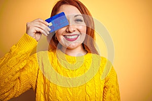 Young beautiful redhead woman holding credit card over yellow isolated background with a happy face standing and smiling with a