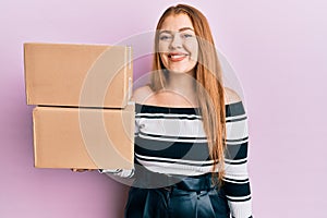 Young beautiful redhead woman holding delivery package looking positive and happy standing and smiling with a confident smile