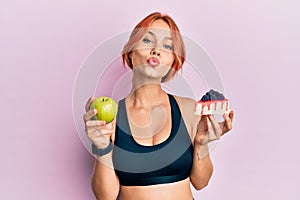 Young beautiful redhead woman holding green apple and cake slice looking at the camera blowing a kiss being lovely and sexy