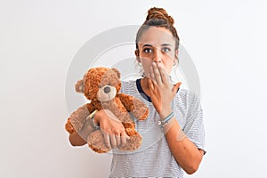 Young beautiful redhead woman holding stuffed teddy bear over isolated background cover mouth with hand shocked with shame for