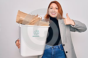 Young beautiful redhead woman recycling holding trash can with cardboard to recycle happy with big smile doing ok sign, thumb up