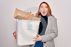 Young beautiful redhead woman recycling holding trash can with cardboard to recycle scared in shock with a surprise face, afraid