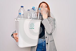 Young beautiful redhead woman recycling holding trash can with plastic bottles to recycle cover mouth with hand shocked with shame