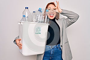 Young beautiful redhead woman recycling holding trash can with plastic bottles to recycle with happy face smiling doing ok sign