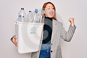 Young beautiful redhead woman recycling holding trash can with plastic bottles to recycle pointing and showing with thumb up to