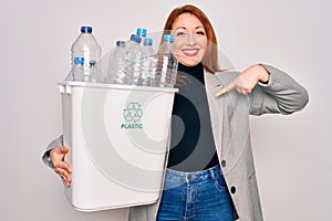 Young beautiful redhead woman recycling holding trash can with plastic bottles to recycle with surprise face pointing finger to