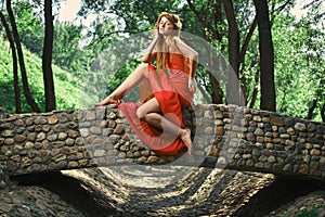 Young beautiful redhead woman in a red dress sits on a stone bridge