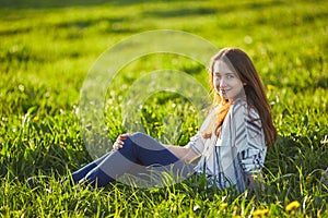 Young beautiful redhead woman sits on a green meadow, looking at camera