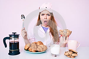 Young beautiful redhead woman sitting on the table having breakfast in shock face, looking skeptical and sarcastic, surprised with