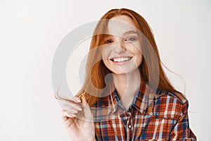 Young beautiful redhead woman smiling with white perfect teeth at camera, touching ginger hair strand and looking happy