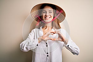 Young beautiful redhead woman wearing asian traditional conical hat over white background smiling in love showing heart symbol and