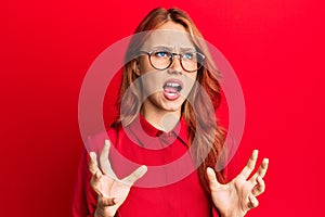 Young beautiful redhead woman wearing casual clothes and glasses over red background crazy and mad shouting and yelling with