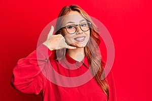 Young beautiful redhead woman wearing casual clothes and glasses over red background smiling doing phone gesture with hand and