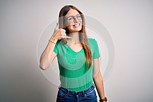 Young beautiful redhead woman wearing casual green t-shirt and glasses over white background smiling doing phone gesture with hand