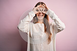 Young beautiful redhead woman wearing casual sweater and glasses over pink background doing ok gesture like binoculars sticking