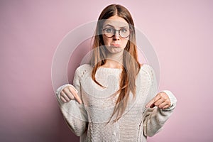 Young beautiful redhead woman wearing casual sweater and glasses over pink background Pointing down looking sad and upset,