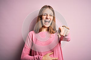 Young beautiful redhead woman wearing casual sweater over isolated pink background laughing at you, pointing finger to the camera