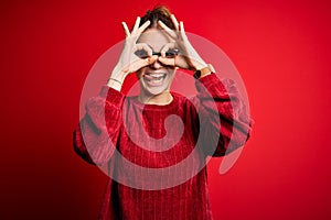 Young beautiful redhead woman wearing casual sweater over isolated red background doing ok gesture like binoculars sticking tongue