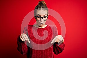 Young beautiful redhead woman wearing casual sweater over isolated red background Pointing down with fingers showing