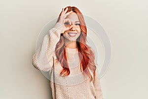 Young beautiful redhead woman wearing casual winter sweater doing ok gesture with hand smiling, eye looking through fingers with