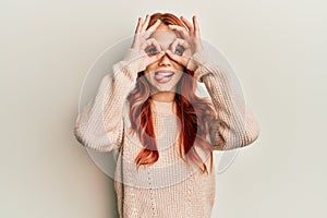 Young beautiful redhead woman wearing casual winter sweater doing ok gesture like binoculars sticking tongue out, eyes looking