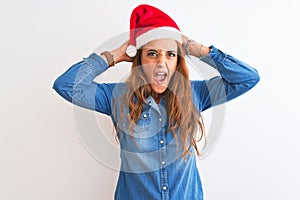 Young beautiful redhead woman wearing christmas hat over isolated background Crazy and scared with hands on head, afraid and