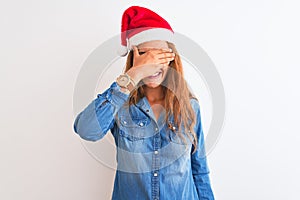 Young beautiful redhead woman wearing christmas hat over isolated background smiling and laughing with hand on face covering eyes