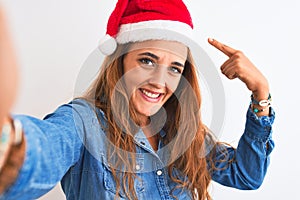 Young beautiful redhead woman wearing christmas hat and taking a selfie very happy pointing with hand and finger