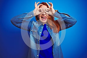 Young beautiful redhead woman wearing denim shirt standing over blue isolated background doing ok gesture like binoculars sticking