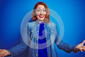 Young beautiful redhead woman wearing denim shirt standing over blue isolated background smiling cheerful with open arms as