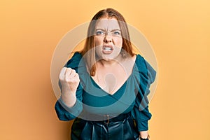 Young beautiful redhead woman wearing elegant and sexy look angry and mad raising fist frustrated and furious while shouting with