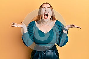 Young beautiful redhead woman wearing elegant and sexy look celebrating mad and crazy for success with arms raised and closed eyes