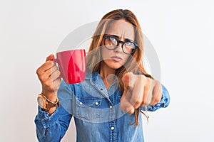 Young beautiful redhead woman wearing glasses drinking cup of coffee over isolated background pointing with finger to the camera