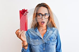 Young beautiful redhead woman wearing glasses and holding birthday present scared in shock with a surprise face, afraid and