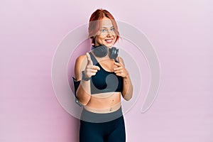 Young beautiful redhead woman wearing gym clothes and using headphones pointing fingers to camera with happy and funny face