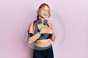 Young beautiful redhead woman wearing gym clothes and using headphones smiling with hands on chest with closed eyes and grateful