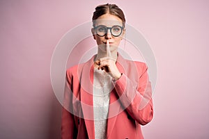 Young beautiful redhead woman wearing jacket and glasses over isolated pink background asking to be quiet with finger on lips