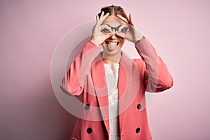 Young beautiful redhead woman wearing jacket and glasses over isolated pink background doing ok gesture like binoculars sticking