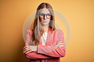 Young beautiful redhead woman wearing jacket and glasses over isolated yellow background skeptic and nervous, disapproving