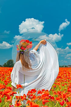 Young beautiful redhead woman in a white shirt with a poppy wreath on her head standing backwards in a poppy field on